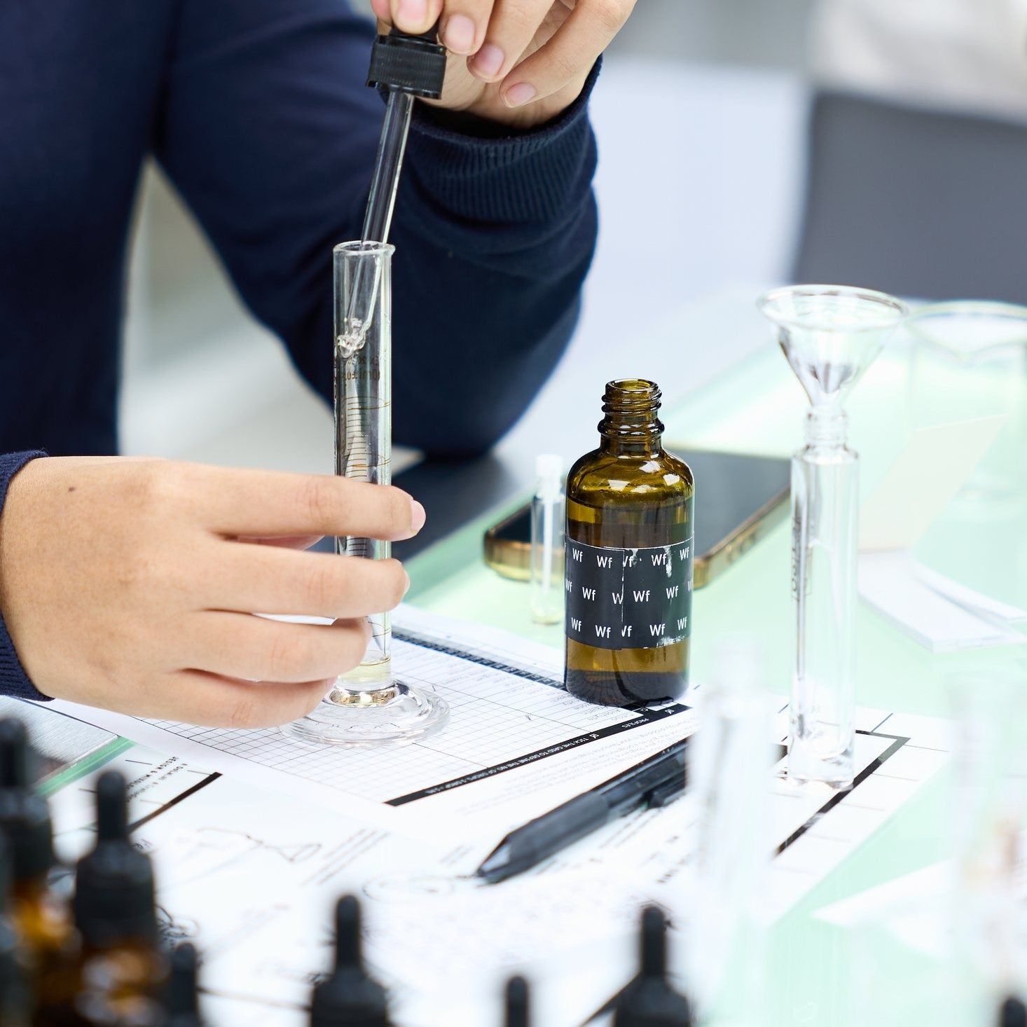 A hand uses a glass pipette to blend perfume at the fragrance-making bench.